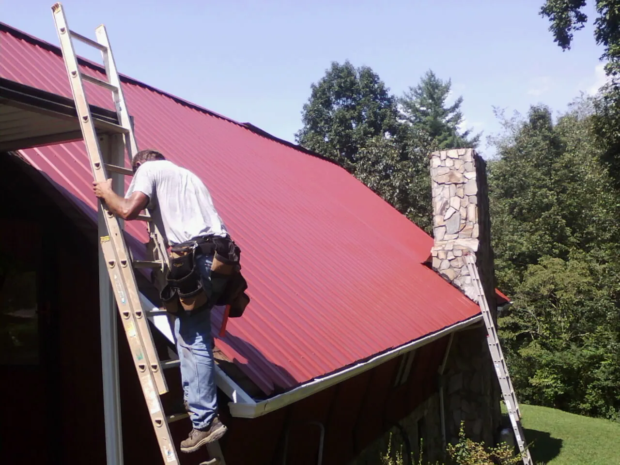 Roofer on a ladder beside a red metal roof and stone chimney — Soffit & Fascia Repair by Roofing Brothers in Pawling