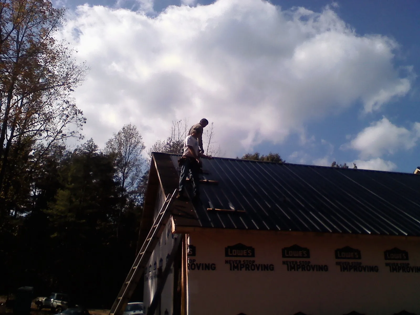 Roofer fastening dark metal panels high against a cloudy sky, from a Roof Inspection job in Pawling